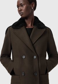 Casaco castanho de dupla fecho com colarinho de pelo sintético preto, grandes botões pretos, tecido texturizado e bolsos laterais.