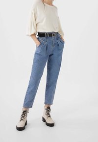 Jersey oversized color beige claro con textura acanalada, combinado con unos vaqueros de mezclilla azules de tiro alto con pliegues. Botas beige con cordones y suelas negras.