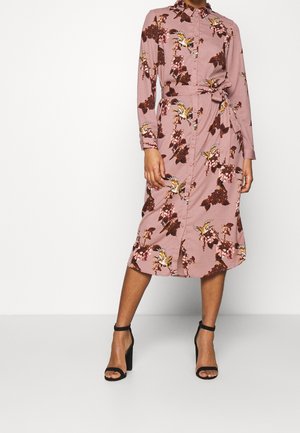 Robe rose à manches longues avec un motif floral en rouge et jaune. Dotée d'une fermeture à boutons et d'une ceinture à la taille. Portée avec des sandales à talons noires.