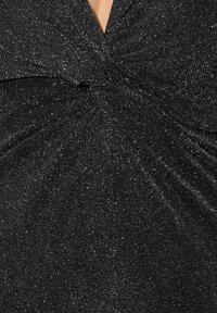 Musta mekko, jossa on tekstuurinen ja kimalteleva kangas. Ominaisuuksiin kuuluu kiertynyt yksityiskohta kaula-aukossa, joka luo V-muotoisen vaikutelman, ja se on istuva malli.