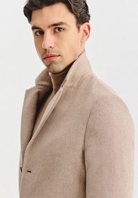 Beżowy wełniany płaszcz z wysokim kołnierzem, dwoma przednimi kieszeniami i dopasowanym krojem. Noszony na jasnobrązowym golfie. Miękka tekstura.
