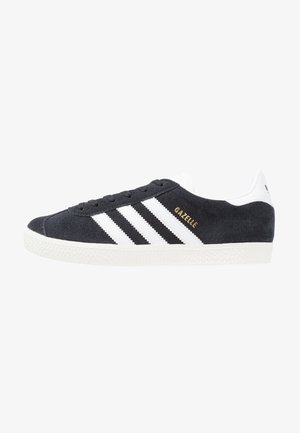 Zapatilla Adidas Gazelle de ante negro con rayas blancas, texto dorado "GAZELLE", suela blanca y lengüeta trasera blanca vista de perfil.