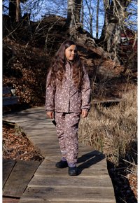 Chaqueta impermeable con estampado floral morado y pantalones a juego, ambos con puños elásticos, usados con zapatos impermeables negros sobre un camino de madera.