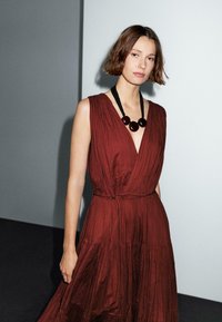 A woman with shoulder-length hair wears a deep V-neck, rust-colored dress and a bold necklace, standing against a minimalist backdrop.