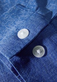 Tessuto blu con motivo testurizzato, caratterizzato da due bottoni di plastica bianchi con un disegno a croce, posizionati su un bordo piegato.