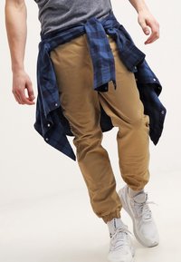 Osoba nosząca beżowe dresowe spodnie, białe trampki, szary t-shirt, z niebieską w kratkę koszulą związaną w talii, idąca do przodu na białym tle.