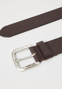 Bruine leren riem met een zilveren metalen gesp. De gesp heeft een gegraveerde "REPLAY"-detail. Glad oppervlak en afgeronde uiteinden met meerdere gaatjes.