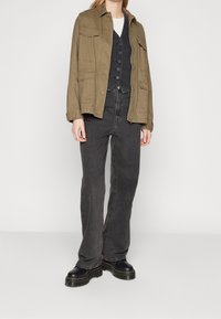 Jasnozielona kurtka z płótna z detalami guzików, warstwowa na czarnym kamizelce i białym t-shircie, zestawiona z czarnymi dzwonami i czarnymi butami.