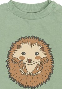 Zelená košeľa s grafickým motívom ježka, s okrúhlym telom, hnedými pichľavými ostňami a svetlou tvárou, na jednofarebnom pozadí.