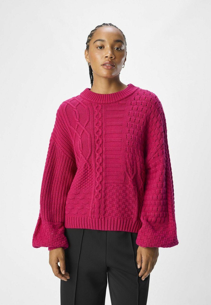 Fuchsia gebreide trui met kabel- en textuurpatronen, brede mouwen en een geribbelde halslijn. Gepaard met zwarte broek.
