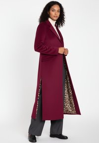 Weinroter langer Mantel mit zwei seitlichen Schlitzen, Kerbkragen und Innenfutter mit Leopardenmuster. Kombiniert mit grauen, weit geschnittenen Hosen und schwarzen Schuhen.
