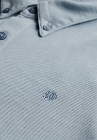 Lichtblauw poloshirt met een kraag en knopen, met een klein geborduurd "JO"-logo nabij de borst.