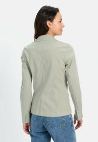 Lichtgroene getailleerde jas met lange mouwen, kraag, naden op de rug en textiel met structuur. Gedragen met blauwe jeans.