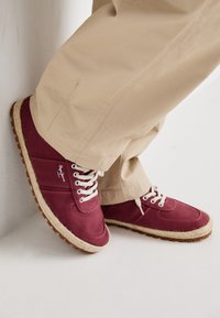 Baskets bordeaux avec des lacets blancs, une tige en toile et une semelle texturée de couleur marron clair avec des détails en jute. Portées avec un pantalon beige.