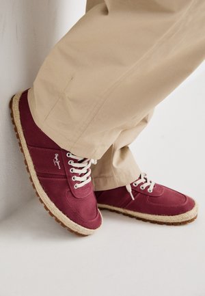 Burgundi színű tornacipő fehér fűzőkkel, vászon felsőrésszel és texturált, világosbarna talppal, juta részletezéssel. Bézs nadrággal viselve.