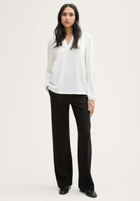 Weiße Bluse mit langen Ärmeln, V-Ausschnitt und lockerer Passform, kombiniert mit schwarzen, weiten Hosen und schwarzen Schuhen.