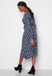 Granatowa sukienka w kwiaty z długimi rękawami, z dopasowanym talią i asymetrycznym dołem. Udekorowana małymi różowymi i białymi kwiatami. Czarne botki za kostkę.