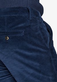Primo piano di una tasca di pantaloni in corduroy blu navy con bottone e cucitura laterale, indossati con un maglione in maglia scura e texturizzata.