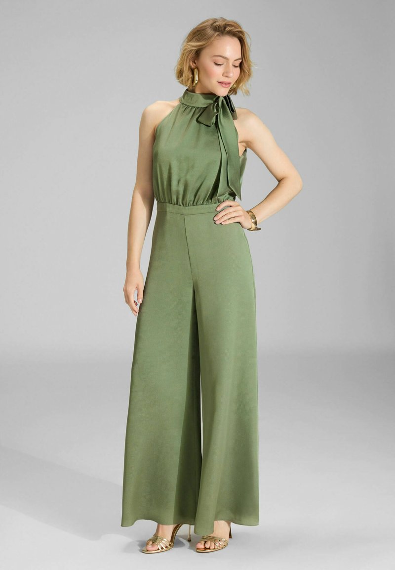 Femme debout portant une combinaison olive verte sans manches avec col halter et jambes larges, des sandales à talons hauts dorées et des boucles d'oreilles anneaux dorées.