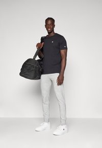Zwarte katoenen t-shirt, grijze sweatpants en witte sneakers. Houdt een zwarte sporttas met gaaspanelen vast. Casual sportkleding.