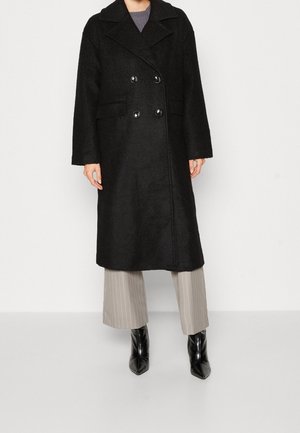 Kobieta w długim, czarnym, dwurzędowym płaszczu, nosząca szare spodnie w cienkie prążki i czarne botki z ostrym noskiem, na jednolitym tle.