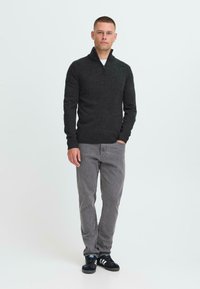 Mann står mot hvit bakgrunn iført mørk glidelåsgenser, grå jeans og svarte Adidas-sneakers med hvite striper.