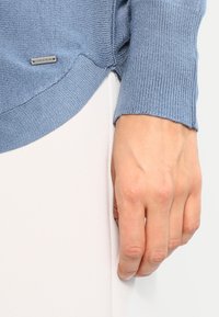 Helles blaues Strickpullover mit gerippten Bündchen und seitlichem Schlitz. Kleines silbernes Markentag am Saum. Die Hand des Modells liegt auf einer weißen Oberfläche.
