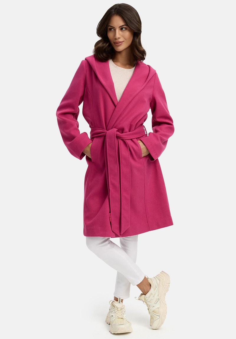 Robe rosa con cappuccio, vita con cintura, maniche lunghe e tasche laterali. Indossato sopra un top leggero e abbinato a pantaloni bianchi e sneaker.