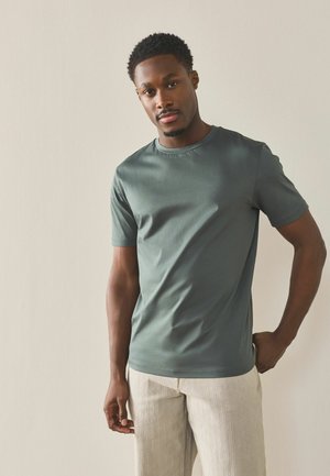 Bărbat cu păr scurt și creț, purtând o cămașă cu mânecă scurtă verde-mușchi și pantaloni bej deschis, stând în fața unui fundal simplu și deschis la culoare.