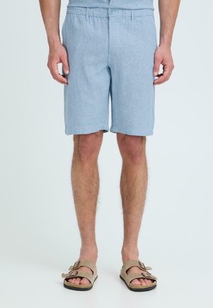 Mann iført lyseblå knelange shorts og beige sandaler med dobbel stropp, stående mot en ensfarget lys bakgrunn.