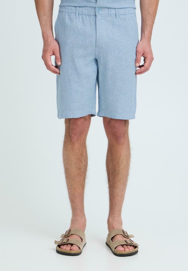 Homme portant un short bleu clair jusqu'aux genoux et des sandales beige à double bride, debout devant un arrière-plan clair uni.