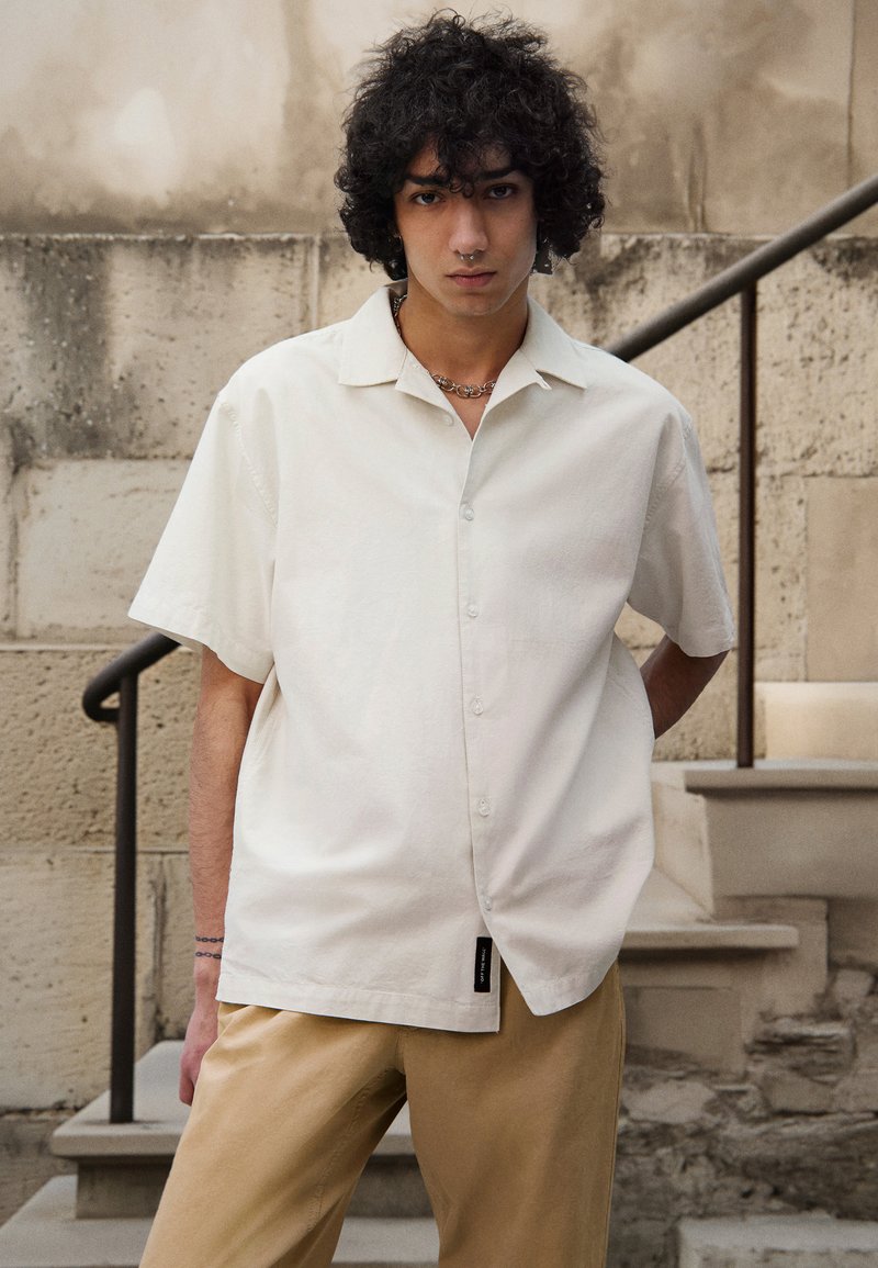 Chemise à manches courtes, col en tissu blanc cassé, avec un devant à boutons et une coupe décontractée ; associée à un pantalon kaki.