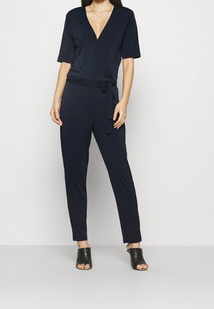 Combinaison bleu marine avec un décolleté en V profond, des manches courtes et une taille nouée. Confectionnée dans un tissu doux, associée à des mules noires à bout ouvert.