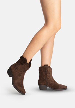 Ben iført mørkebrune ruskind midt på læggen cowboystøvler med sidelås, lav lagdelt hæl og dekorative syninger på en lys baggrund.