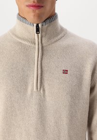 Beżowy sweter zapinany na zamek z prążkowanym szarym kołnierzem. Posiada mały emblemat z flagą Norwegii w kolorze czerwonym i niebieskim na klatce piersiowej. Miękka, dziana faktura.