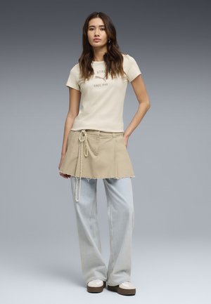 Joven con cabello largo que lleva una camiseta beige ajustada con gráfico, minifalda kaki con cadena de perlas colgante, jeans sueltos azul claro y zapatillas.