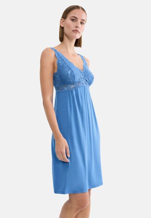 Kobieta w niebieskiej sukience bez rękawów z koronkowymi zdobieniami na biuście i ramiączkach, stojąca na tle jednolitego jasnego tła.