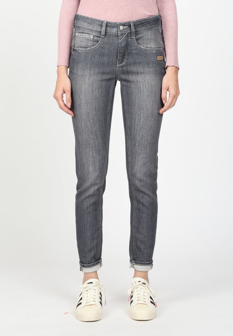 Femme portant un jean slim gris retroussé aux chevilles, un haut à manches longues côtelé rose et des baskets blanches avec des bandes noires.