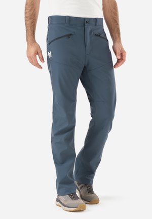 Mann trägt blaue Outdoor-Hose mit Reißverschlusstaschen und graue Wanderschuhe und steht vor weißem Hintergrund.