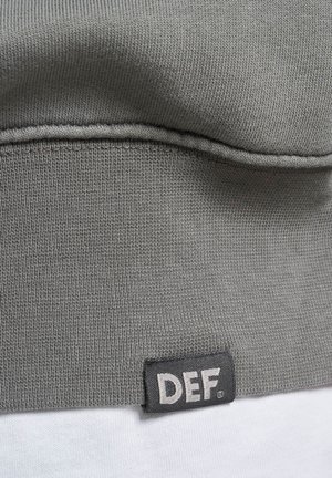 Grijs sweatshirtmateriaal met een geribbeld patroon. Heeft een zwart etiket met witte tekst "DEF" aan de onderzoom. Glad oppervlak en een eenvoudig ontwerp.