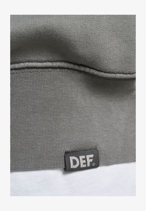 Grijs sweatshirtmateriaal met een geribbeld patroon. Heeft een zwart etiket met witte tekst "DEF" aan de onderzoom. Glad oppervlak en een eenvoudig ontwerp.
