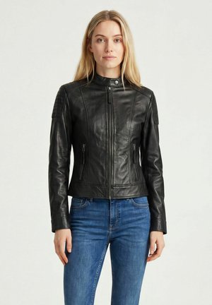 Junge Frau mit langen blonden Haaren, die eine taillierte schwarze Lederjacke und blaue Jeans trägt, steht vor einem schlichten hellen Hintergrund.