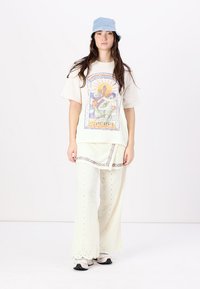 T-shirt graphique blanc avec un design multicolore ; superposé sur une tunique crème et un pantalon large avec des détails en œillets. Chapeau seau bleu.