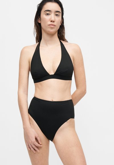 Γυναίκα με βρεγμένα μαλλιά, που φορεί μαύρο μαγιό τοπ halter και μαγιό σλιπ με ψηλή μέση, σε απλό λευκό φόντο.