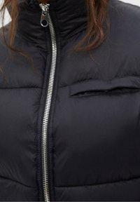 Chaleco acolchado negro con cuello alto, cremallera plateada y un pequeño bolsillo horizontal en el pecho. Textura suave con secciones acolchadas.