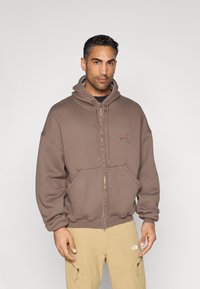 Sweatshirt castanha com fecho de correr, capuz com cordão, bolsos frontais e logótipo bordado. Tecido texturizado; corte solto, estilo descontraído.