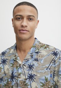Cămașă cu mânecă scurtă, butonată, de culoare maron deschis, cu modele florale și palmieri în nuanțe de albastru și verde; prezintă ilustrații în stil vintage ale unor figuri.