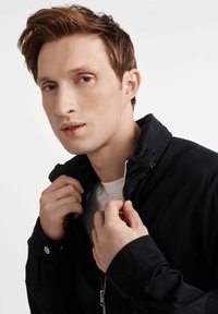 Czarna kurtka zapinana na suwak z wysokim kołnierzem, gładką teksturą i srebrnym suwakiem. Model trzyma kołnierz, ma na sobie białą koszulkę.