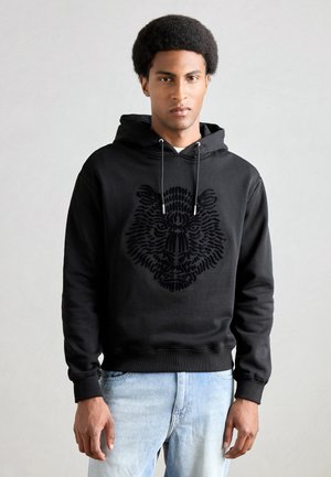 Black hooded sweatshirt featuring an embossed lion design. Made of soft material with a relaxed fit and ribbed cuffs. Paired with light jeans.