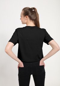 Černé bavlněné tričko s krátkým rukávem, kulatým výstřihem, hladkou texturou, nošené s černými džíny, viděné zezadu.